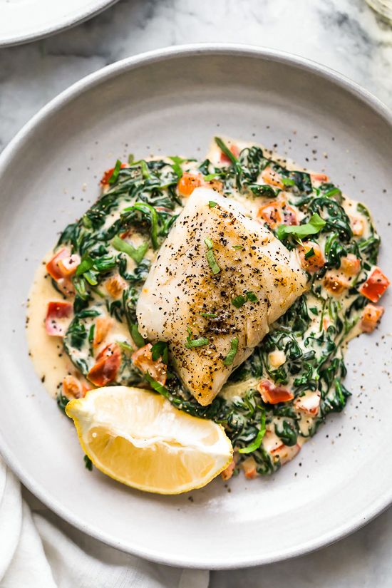 a white plate topped with fish covered in spinach and lemon wedges next to a slice of lemon