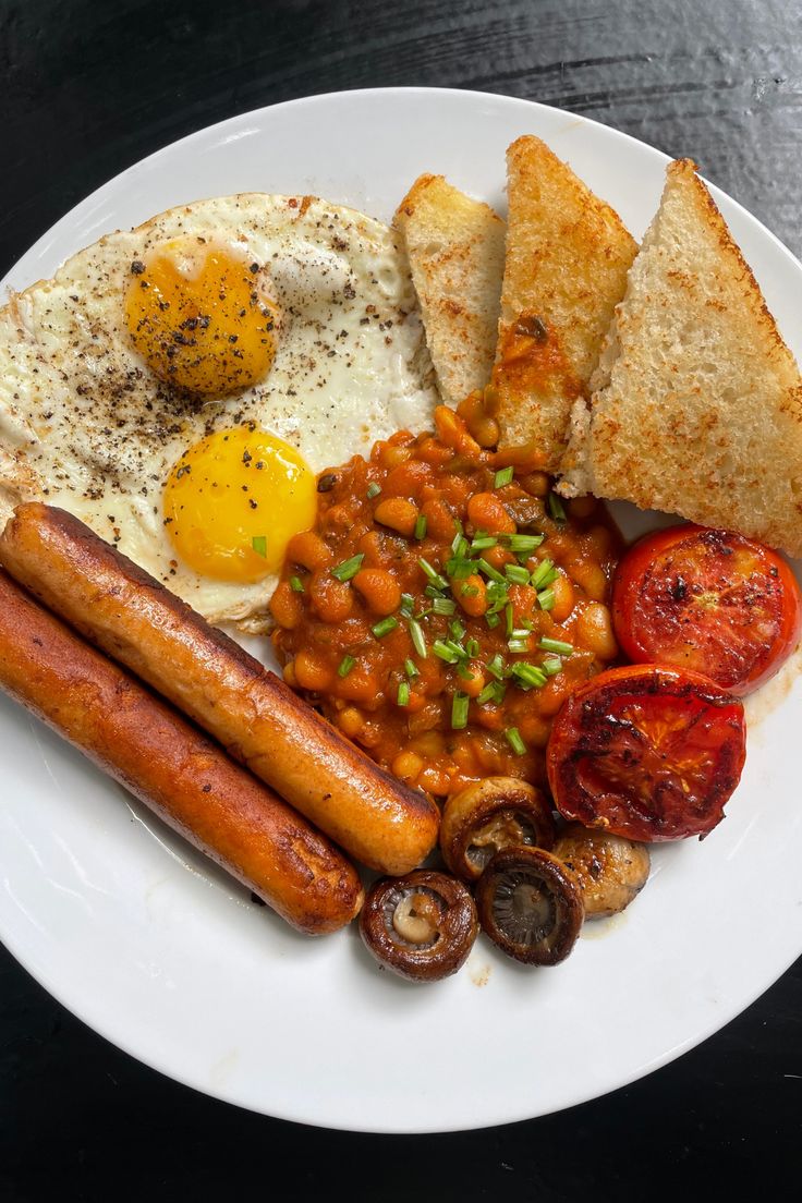 a white plate topped with eggs, sausages and beans next to toasted bread