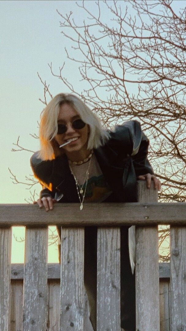 a woman with blonde hair and sunglasses leaning on a wooden fence looking at the camera