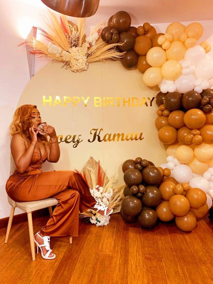 a woman sitting on a chair in front of a birthday balloon arch and talking on the phone