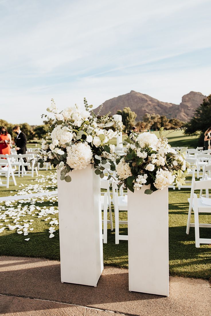 two tall white vases filled with flowers on top of a grass covered field next to chairs