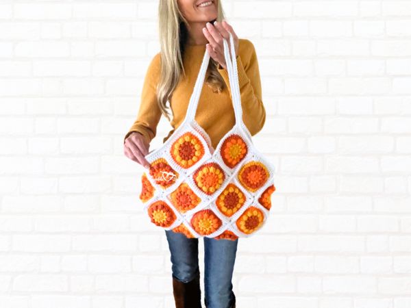 a woman holding a crocheted bag in front of a white brick wall,