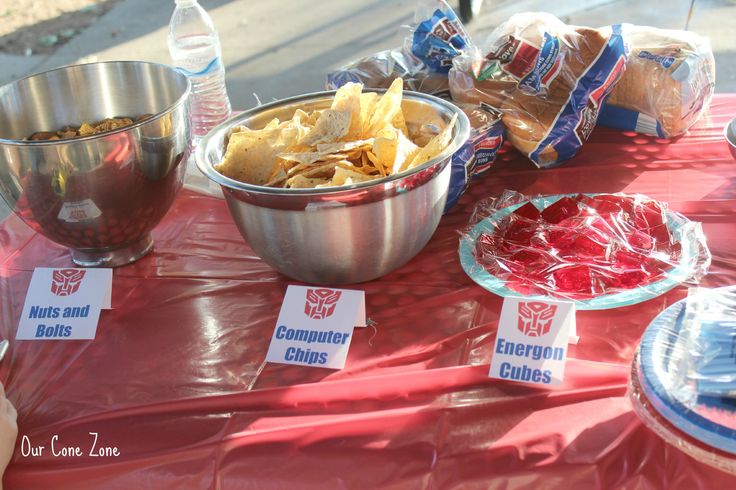 a red table topped with bowls and plates filled with food next to bags of chips