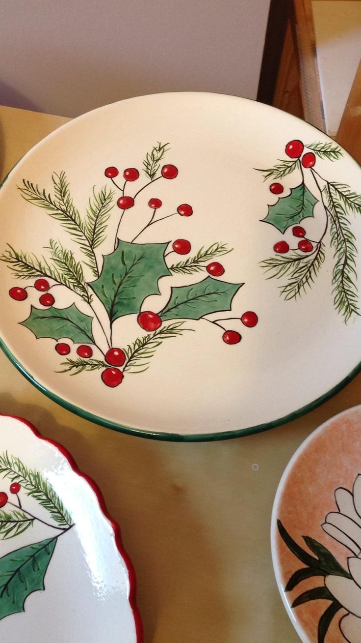 three plates with holly designs on them sitting on a table next to another plate that has flowers and leaves painted on it