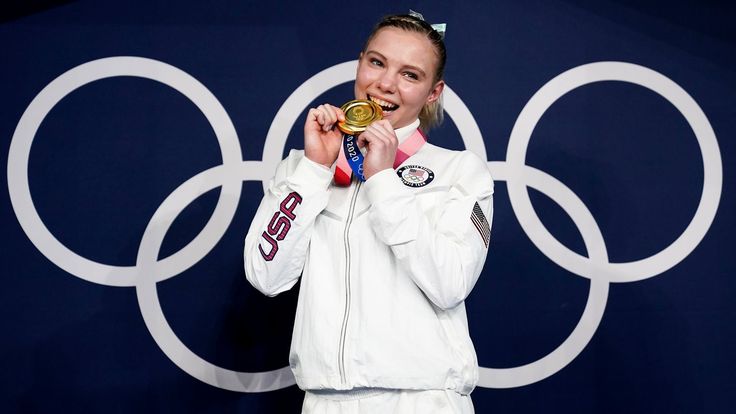 a woman holding a gold medal in front of a blue and white wall with the olympic rings behind her