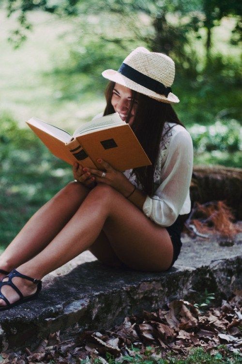 a young woman sitting on the ground reading a book while wearing sandals and a hat