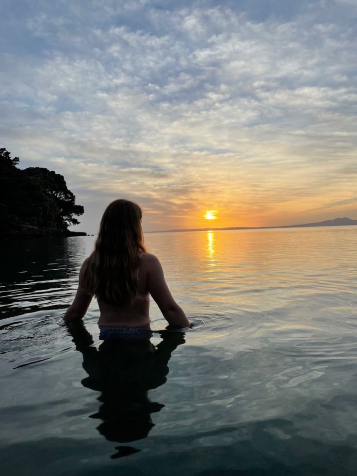 a woman is sitting in the water watching the sun go down over the ocean with her back turned to the camera