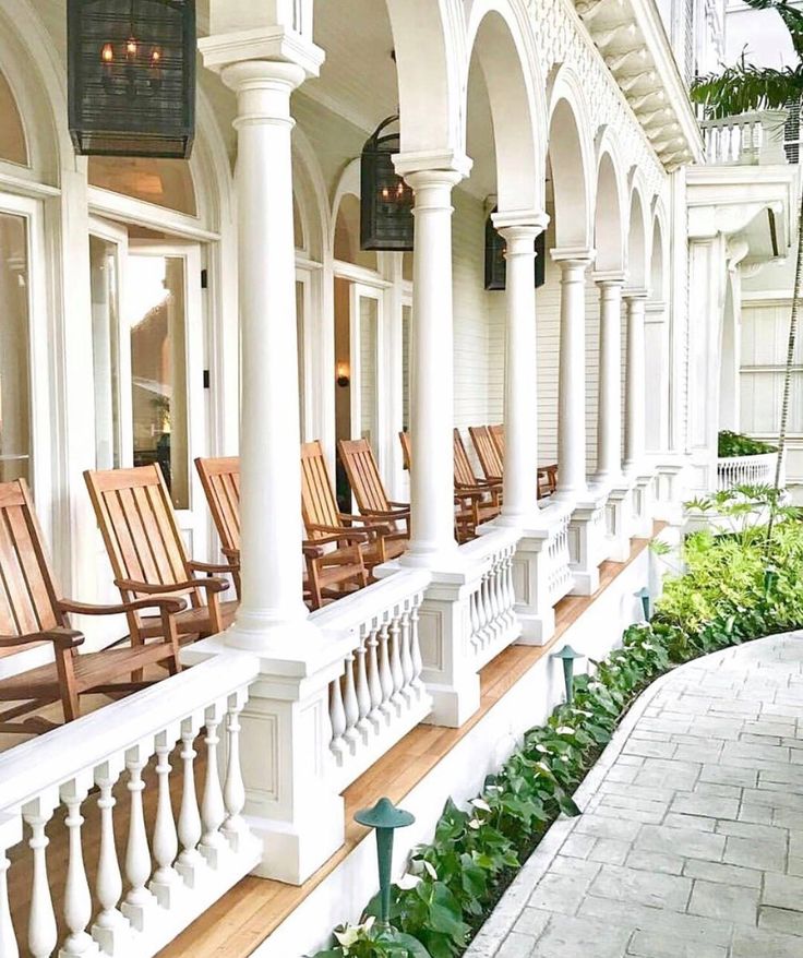 several wooden chairs lined up on the front porch of a white building with columns and windows