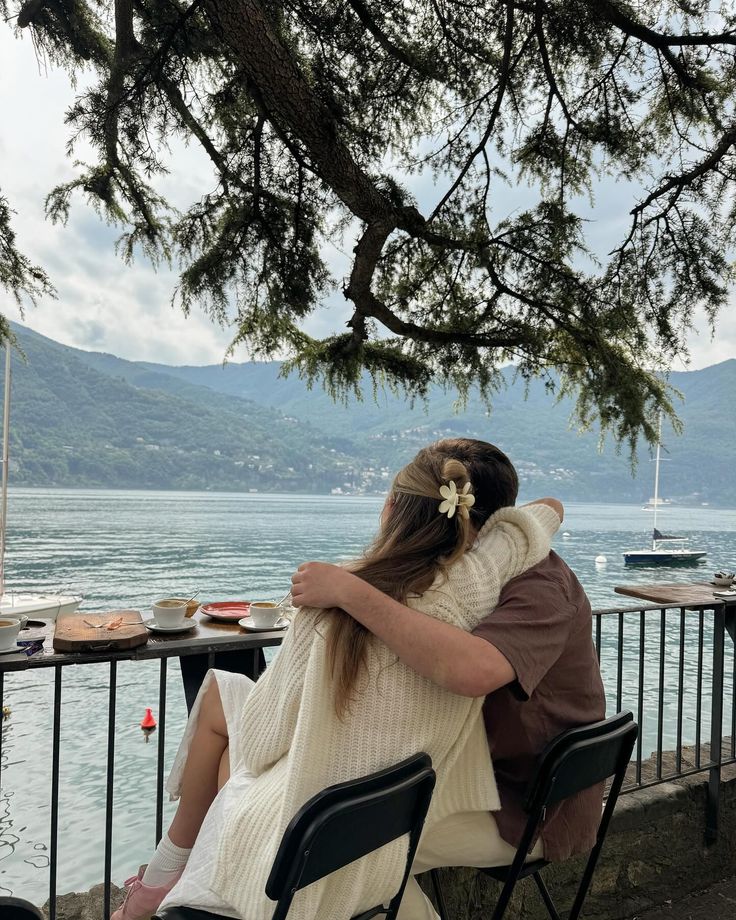 two people are sitting on a bench by the water and one person is hugging her