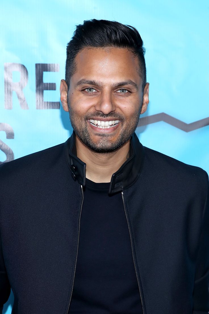 a smiling man in a black jacket and black shirt smiles at the camera while standing in front of a blue background