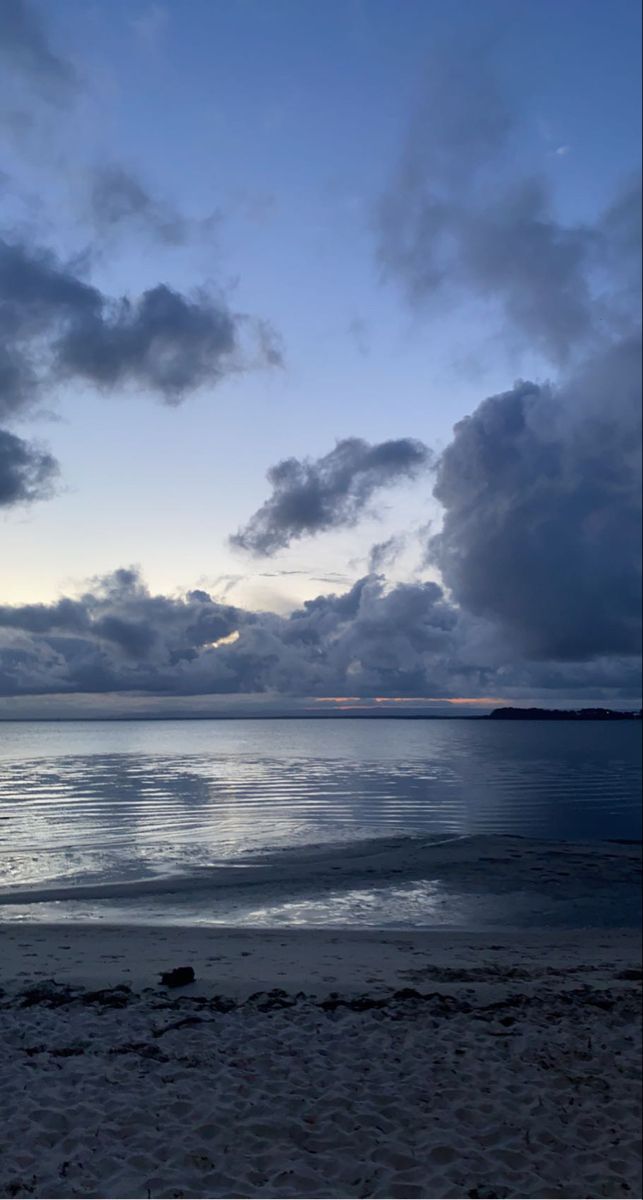 the sky is filled with dark clouds over the water at dusk on an empty beach