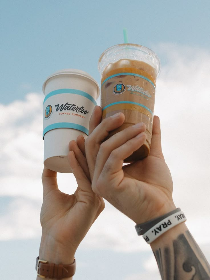 two people holding up coffee cups in front of a blue sky with clouds behind them