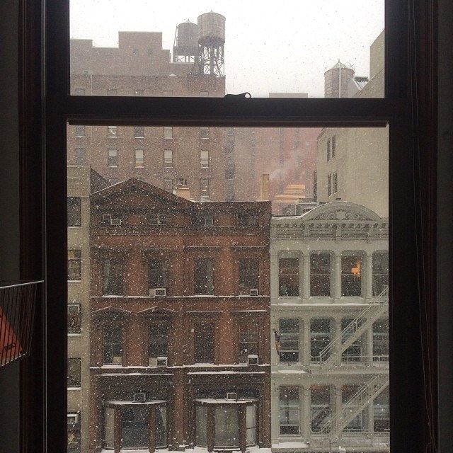 a window with snow on the ground and buildings in the background, looking out at an urban area