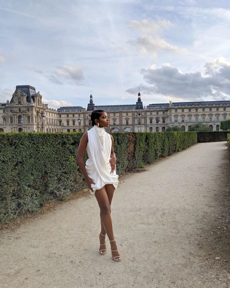 a woman standing in front of a large building with hedges on the sides and a walkway leading to it