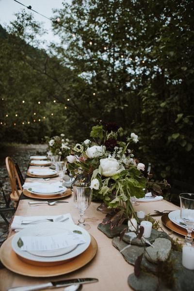 an outdoor table set with place settings and flowers in vases on the table, surrounded by greenery