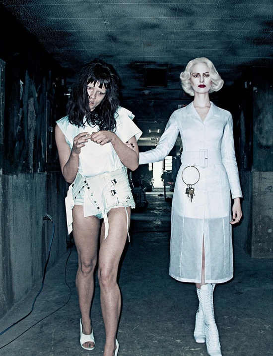 two women dressed in white walking down a dark hallway together, one holding the other's hand