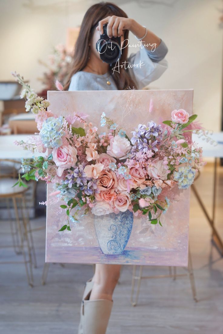 a woman is holding up a painting with flowers in it and she is taking a picture