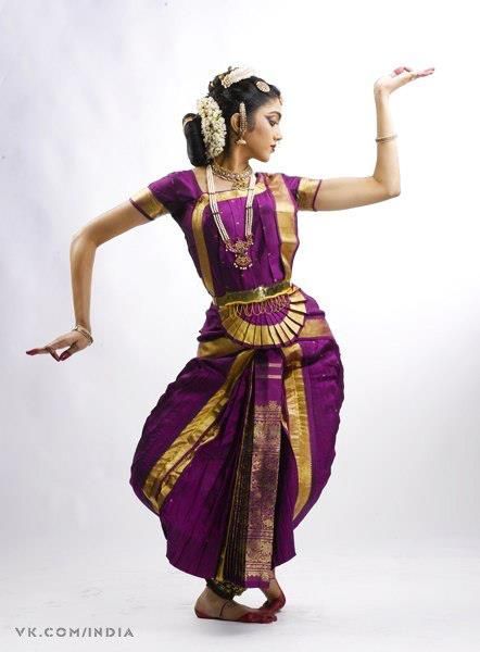 a woman in a purple and gold sari dancing with her arms spread out to the side