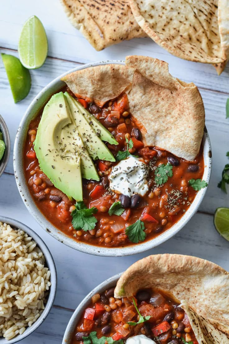 two bowls filled with chili and beans next to tortilla chips, avocado and sour cream