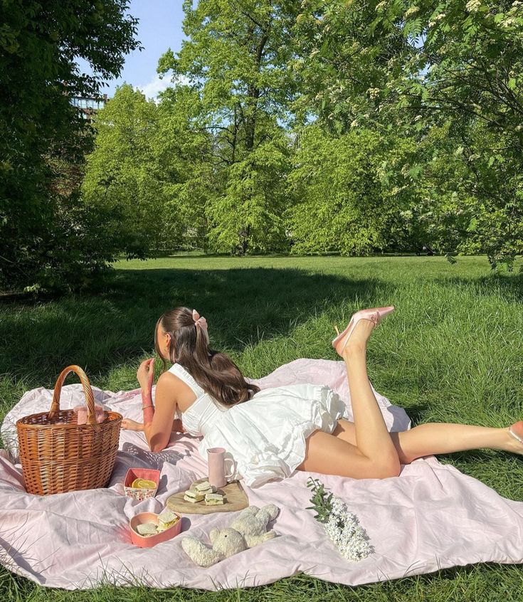 a woman laying on top of a pink blanket next to a picnic basket filled with food