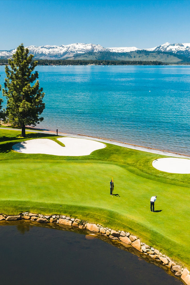 two people are playing golf on the green by the water with mountains in the background