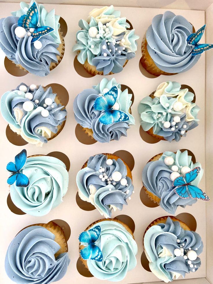 twelve cupcakes with blue frosting and flowers in a box on a table