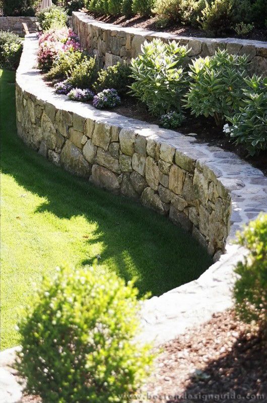 a stone wall and landscaping area in the middle of a yard with green grass, shrubs and flowers