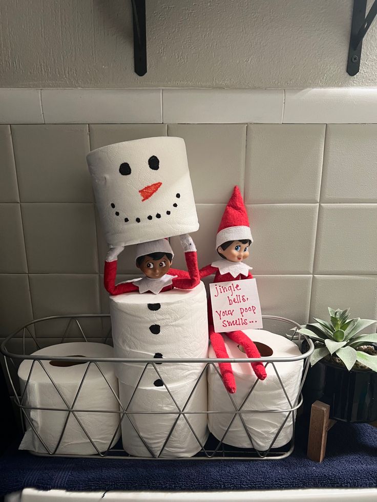 two elfs are sitting on top of toilet paper in the bathroom and one is wearing a snowman hat