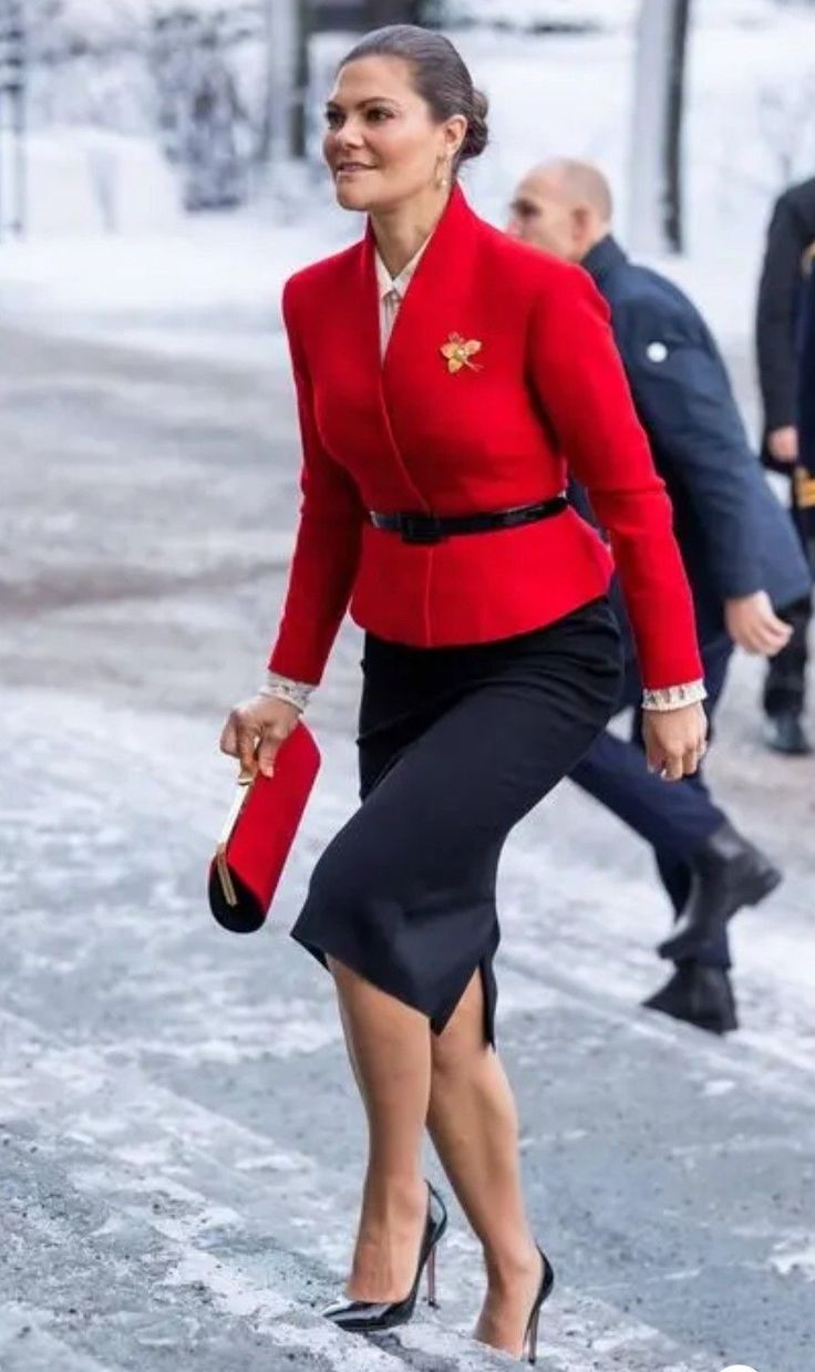 a woman in a red jacket and black skirt is walking through the snow wearing high heels