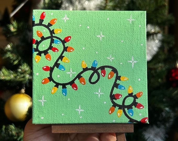 a hand holding up a small square painting with christmas lights on it