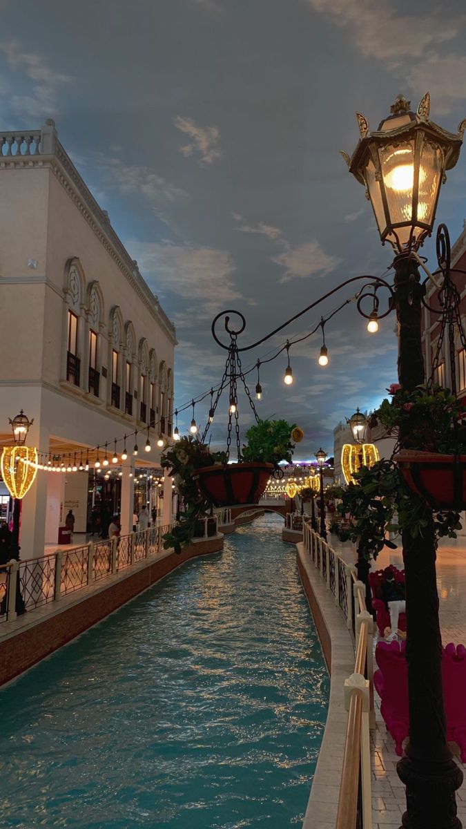 a street light next to a canal in a shopping district at dusk with lights strung from the buildings