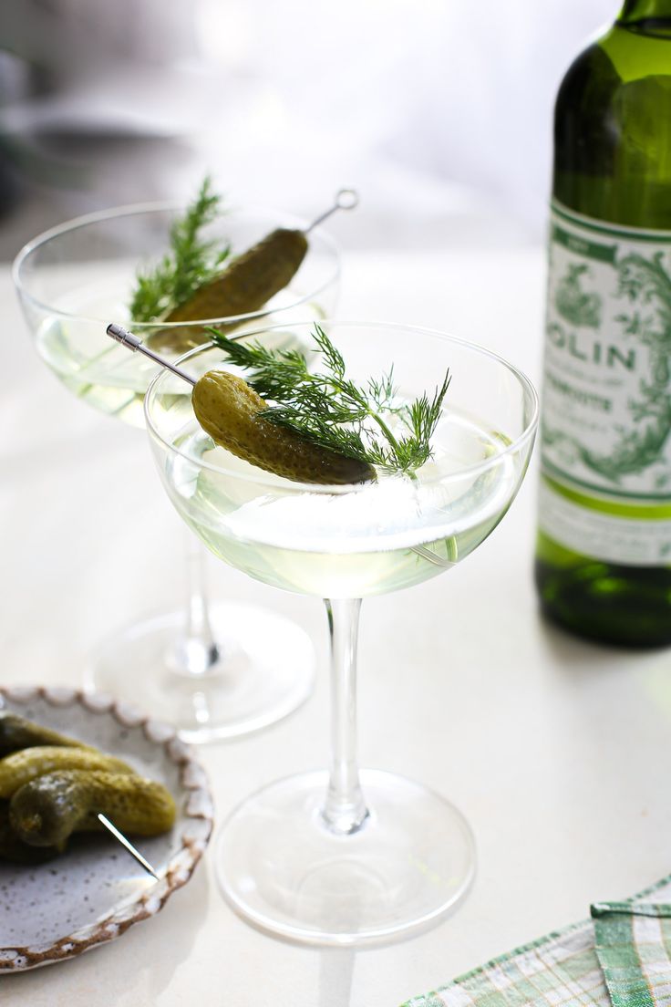 two glasses filled with olives and pickles on a table next to a bottle