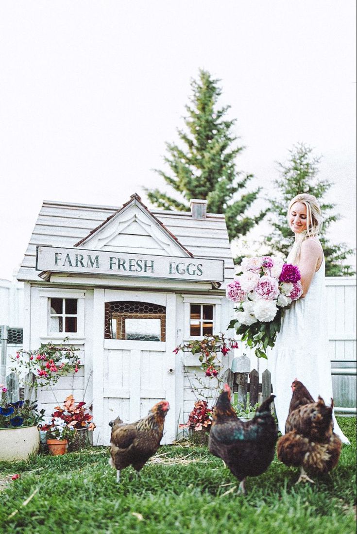 a woman is standing in the grass with chickens and holding a bunch of pink flowers