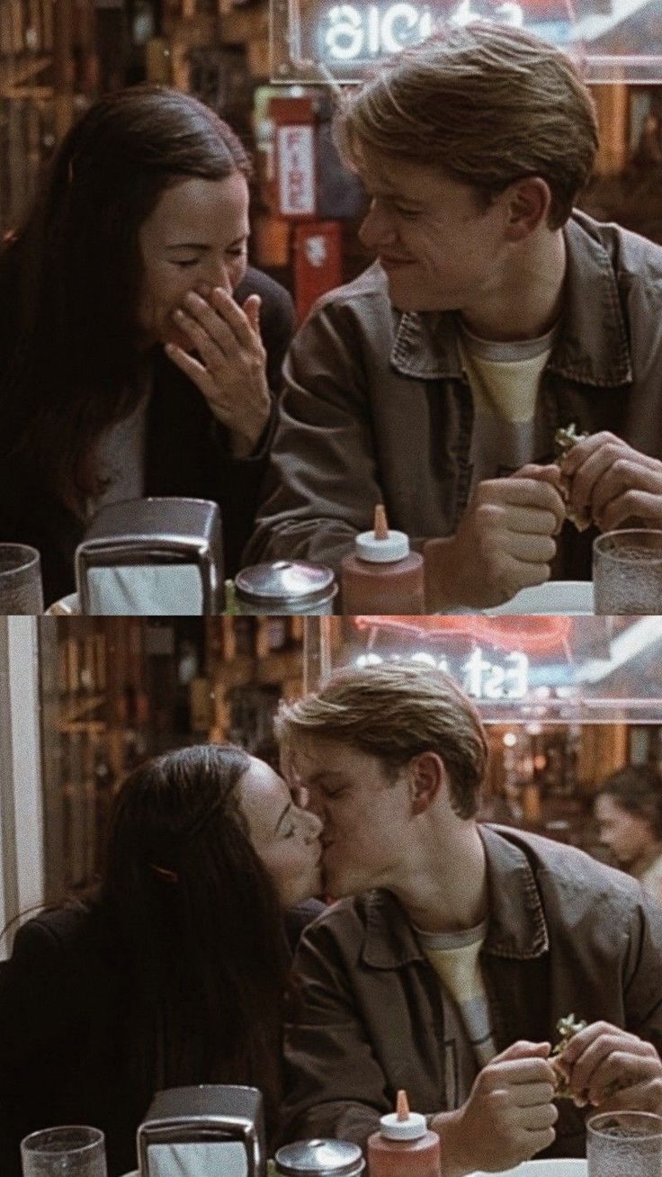 two people sitting at a table and one is kissing the other's face with food in front of them