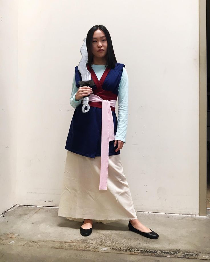 a woman standing in front of a white wall wearing a blue and pink outfit with a red sash around her neck