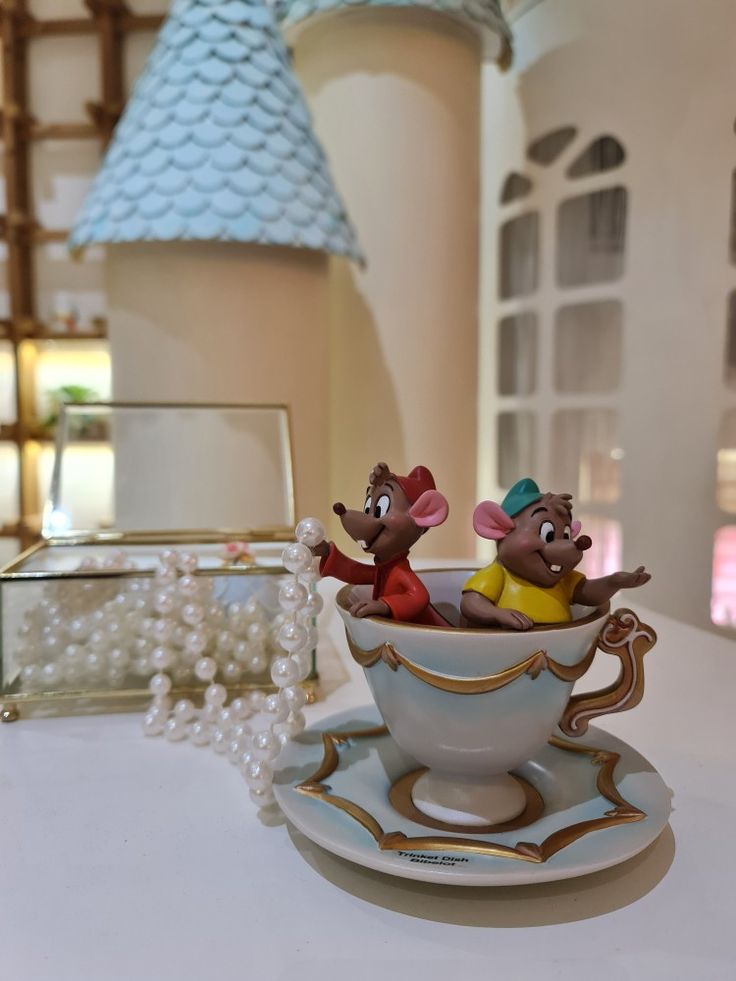 two mouse figurines sitting in a teacup on a table with pearls around it