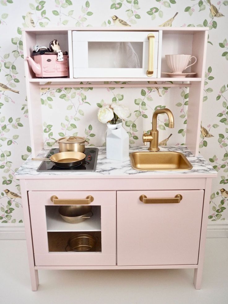 a toy kitchen with pink cabinets and gold accessories on the counter top, along with white wallpaper