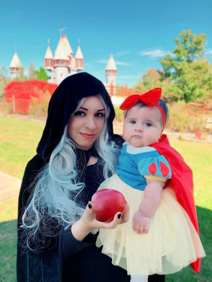 a woman holding a baby dressed as snow white and an apple in front of a castle