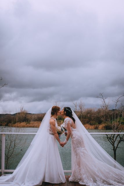 two women in wedding dresses standing on a dock next to the water and holding hands