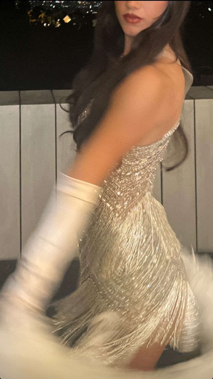 a woman in a silver dress and white gloves is posing for the camera with city lights behind her