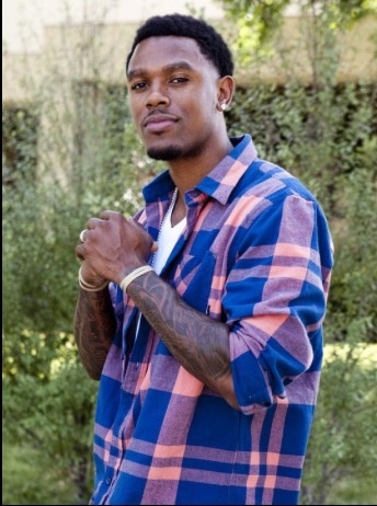 a man with tattoos standing in front of some bushes and trees, holding his hands together