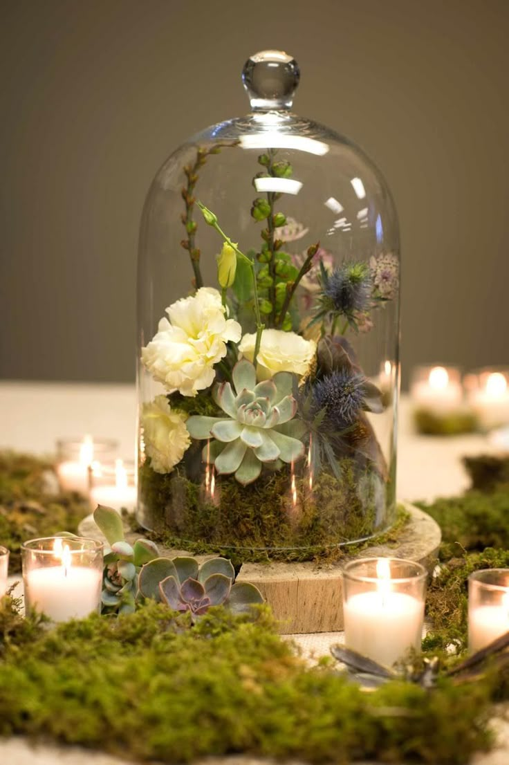 a glass clochel with flowers and greenery on it is surrounded by candles