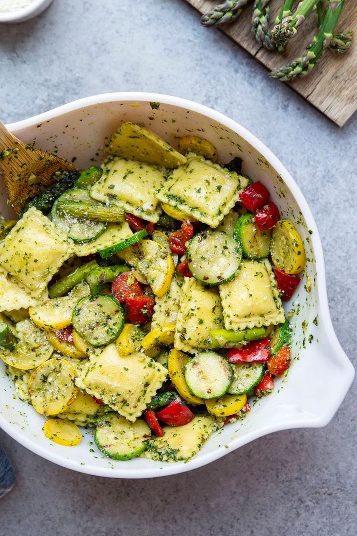 a white bowl filled with zucchini, tomatoes and other veggies next to a cutting board