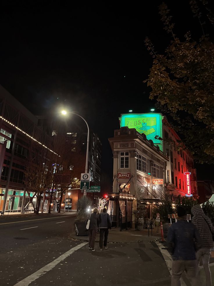 people are walking down the street in front of some buildings at night with lights on