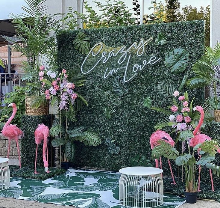 some pink flamingos are standing in front of a green wall with plants and flowers