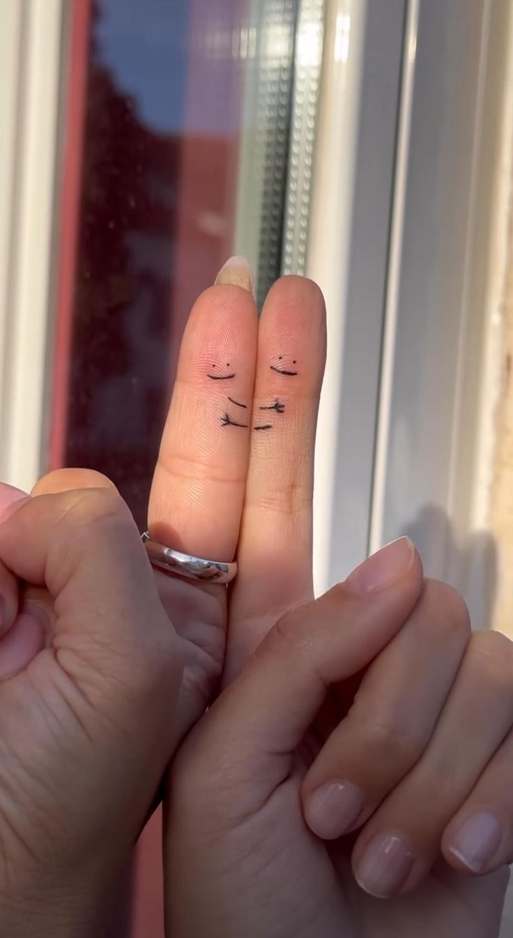 two fingers with small tattoos on them are touching each other's foreheads in front of a window