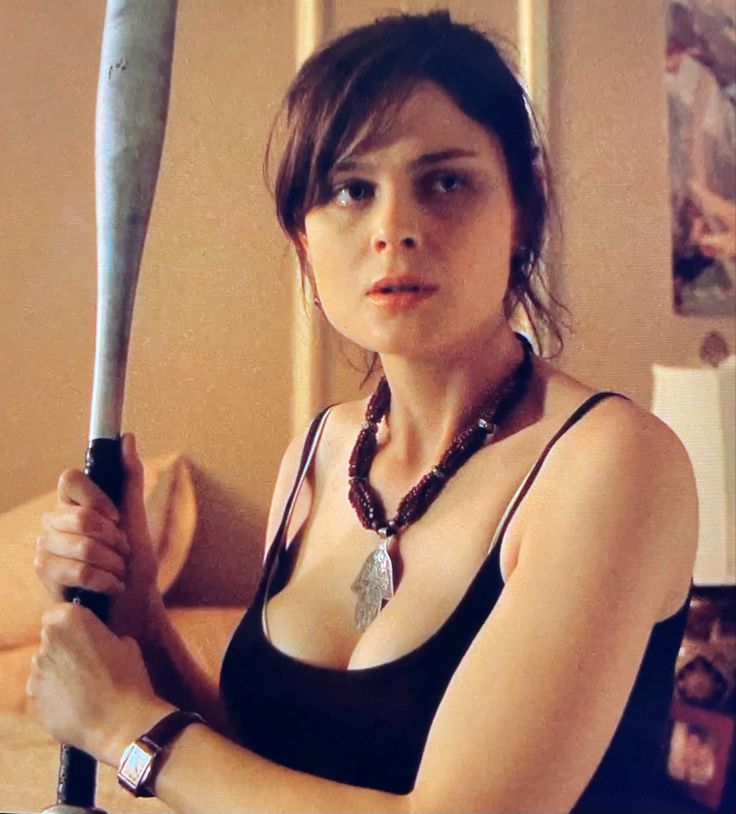 a woman holding a large knife in her right hand and looking at the camera while sitting on a bed