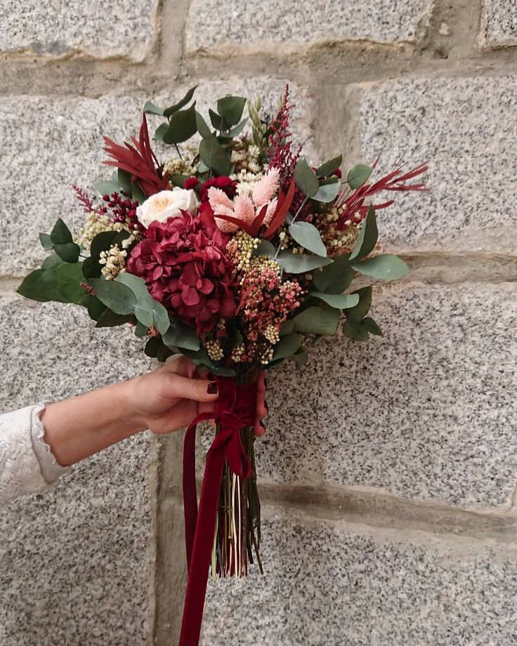 a woman holding a bouquet of flowers against a stone wall with red ribbon tied around it