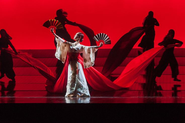 a woman in white and red dress holding a fan on stage with other dancers behind her