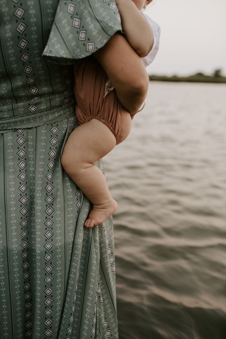 a woman holding a baby in her arms near the water's edge, while wearing a dress